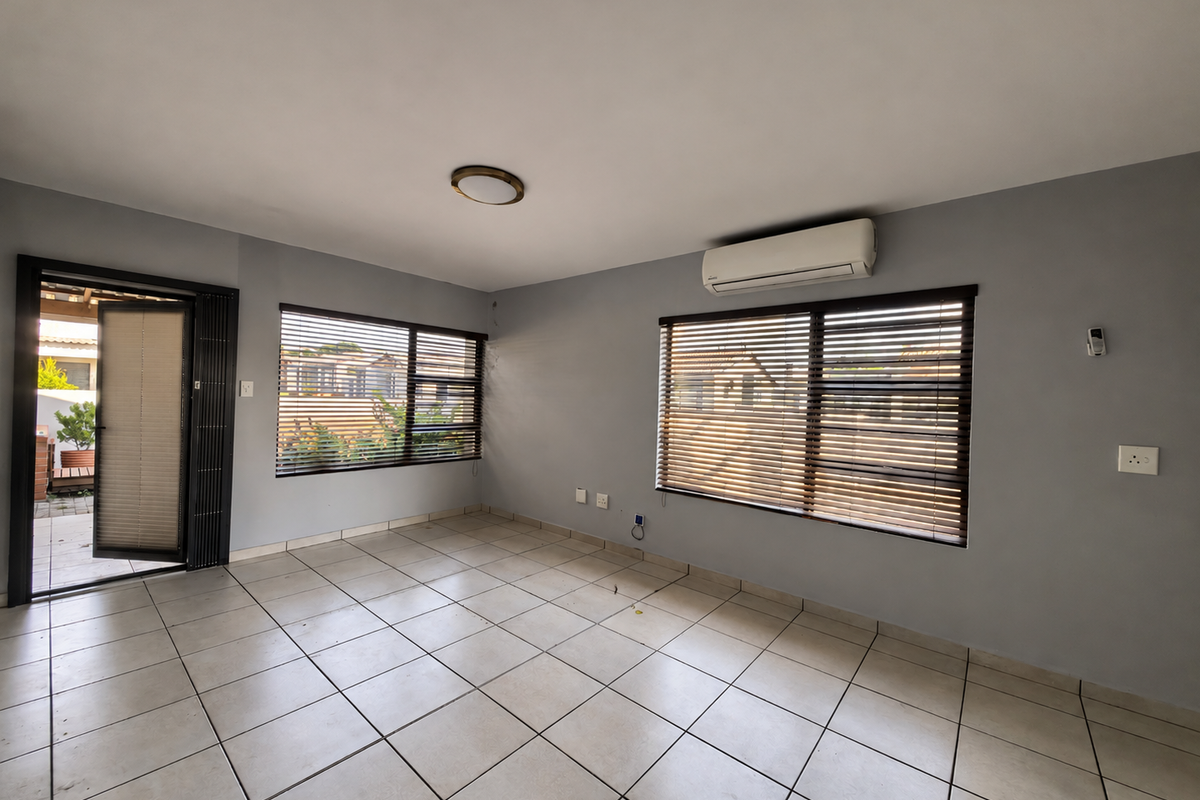 An empty, well-lit living room featuring neutral tiled flooring, grey walls, a wall-mounted air conditioning unit, and venetian blinds on the windows.