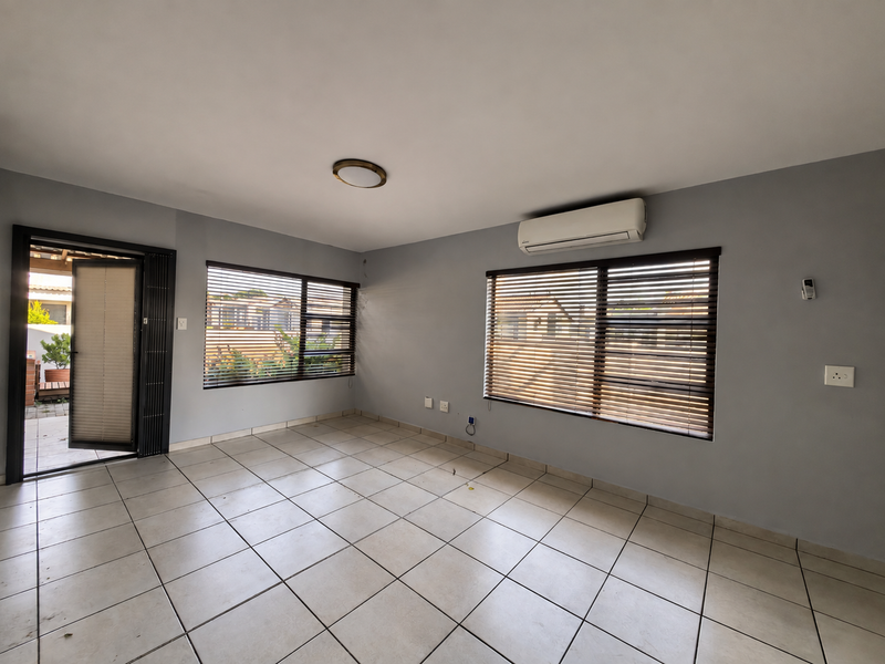 An empty, well-lit living room featuring neutral tiled flooring, grey walls, a wall-mounted air conditioning unit, and venetian blinds on the windows.