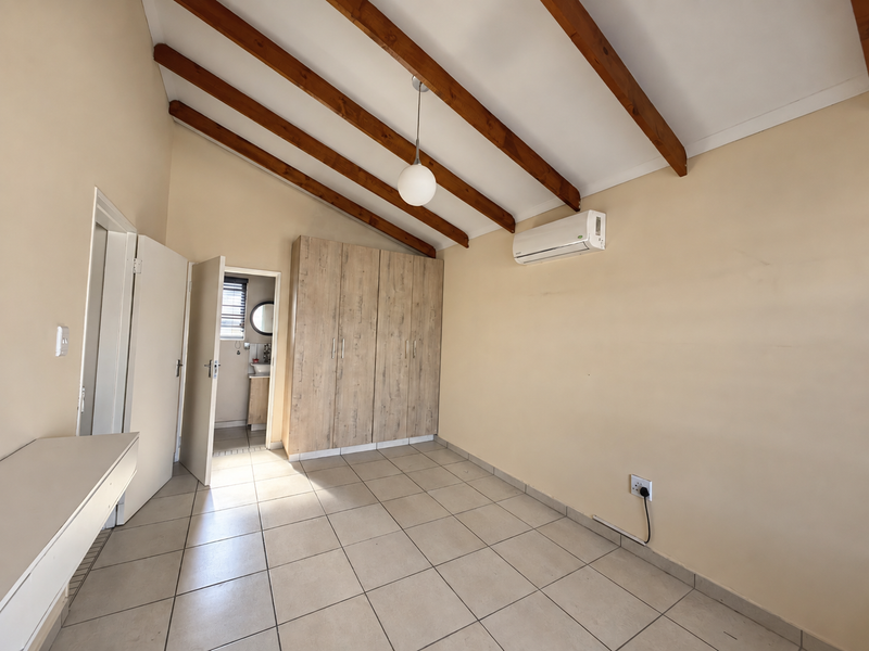 A bright, empty bedroom featuring exposed timber ceiling beams, built-in cupboards, tiled flooring, and a wall-mounted air conditioning unit.
