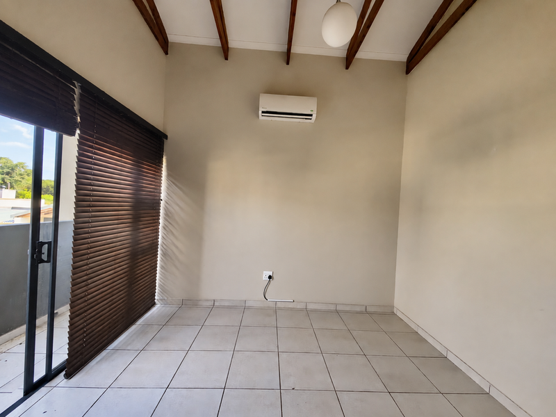 An empty living room featuring exposed timber ceiling beams, tiled flooring, a wall-mounted air conditioner, and a sliding glass door with venetian blinds.
