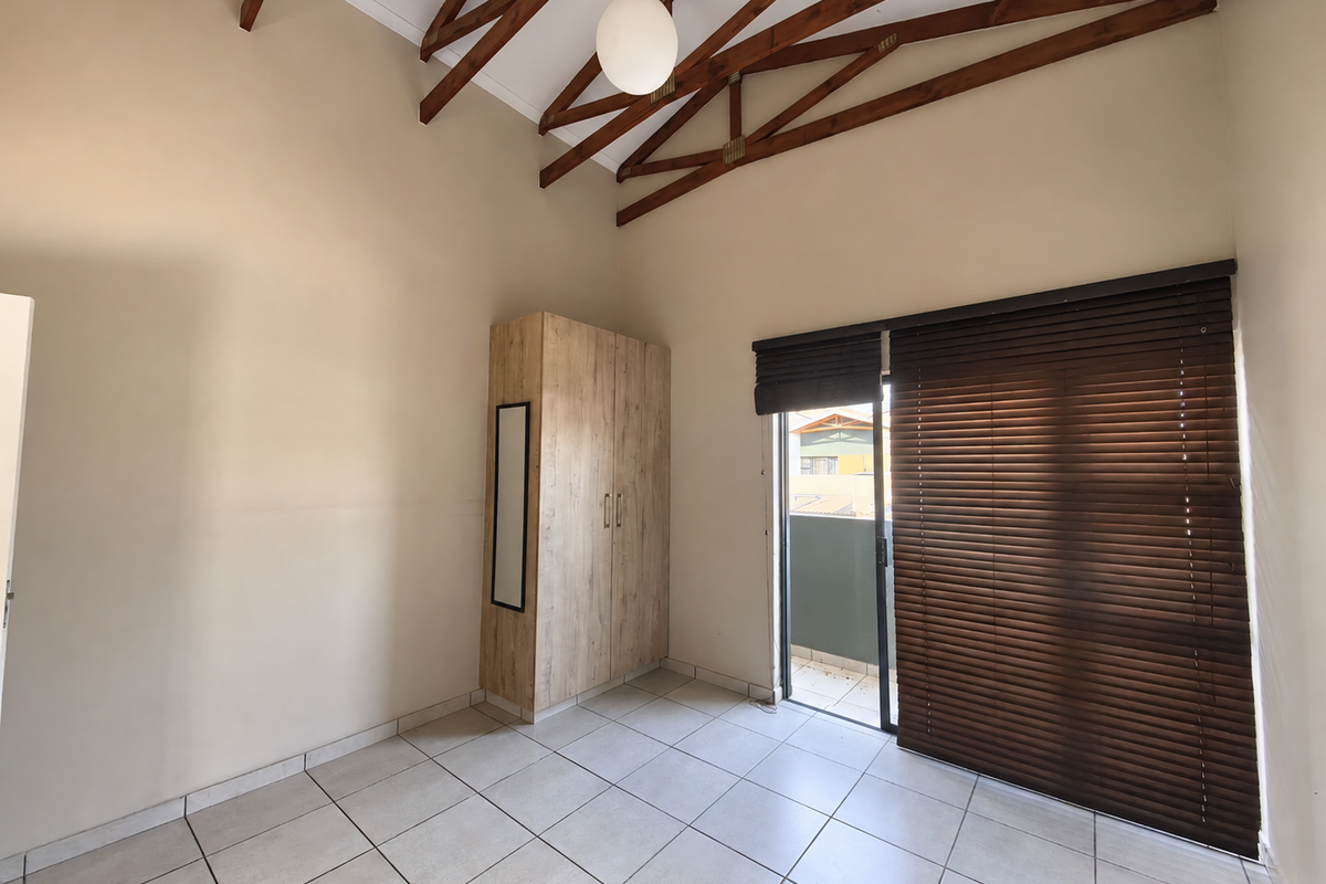 A bright, empty bedroom featuring high vaulted ceilings with exposed timber trusses, tiled flooring, a built-in wardrobe, and direct access to a balcony.