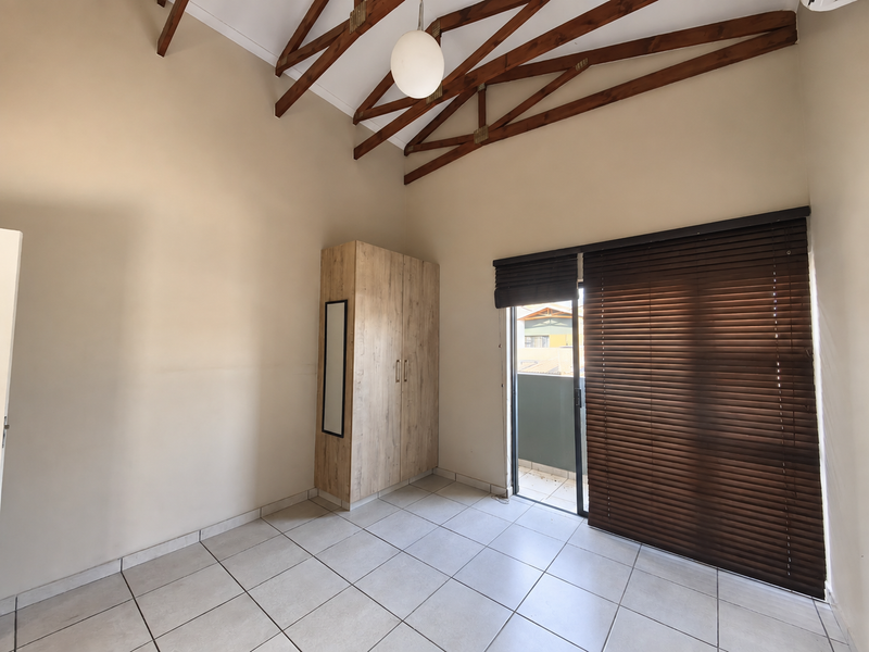 A bright, empty bedroom featuring high vaulted ceilings with exposed timber trusses, tiled flooring, a built-in wardrobe, and direct access to a balcony.