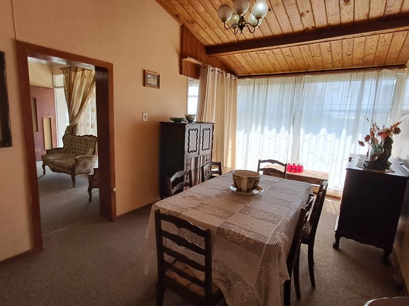 Dining room featuring an exposed timber ceiling, large window for natural light, and carpeted flooring, offering a dated but functional space.