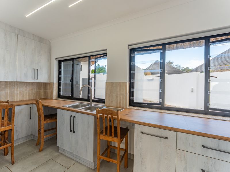 A functional kitchen featuring light wood-grain cabinetry, wooden countertops, a double sink, and tiled splashbacks with natural light from louvre windows.