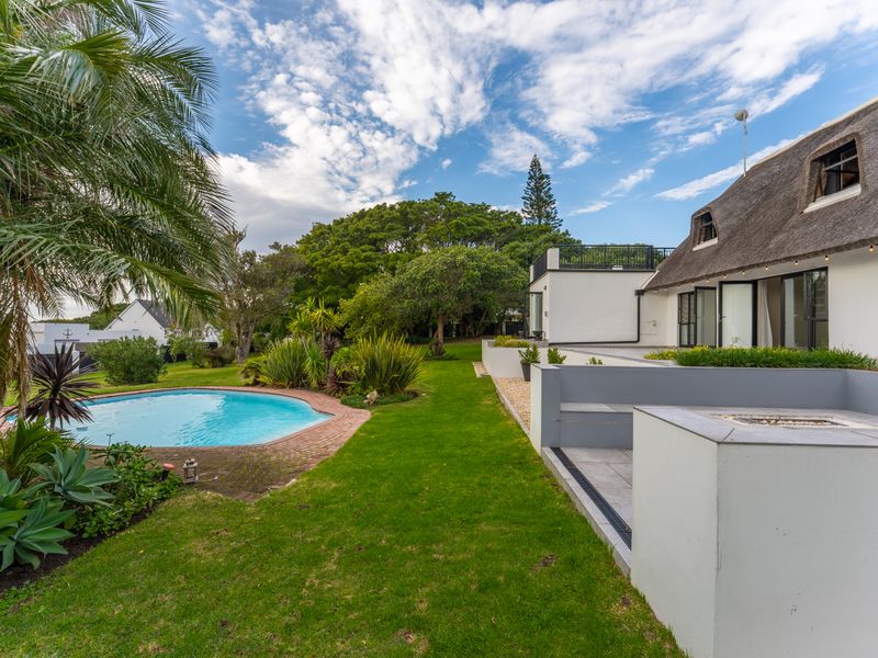 A well-maintained rear garden featuring a swimming pool, a paved patio area with a built-in fire pit, and a house with a traditional thatched roof.