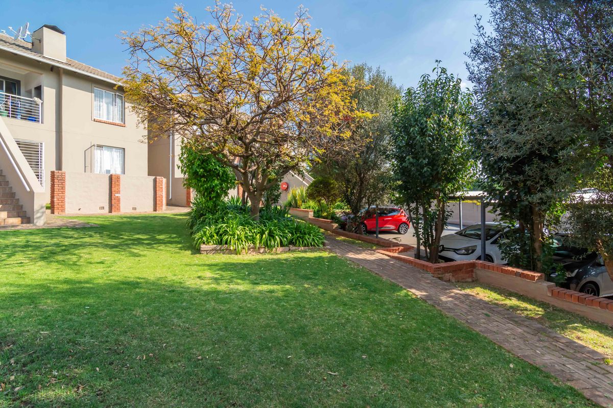 A well-maintained communal garden area featuring a brick pathway, established trees, and a view of the townhouse exterior with balcony access.