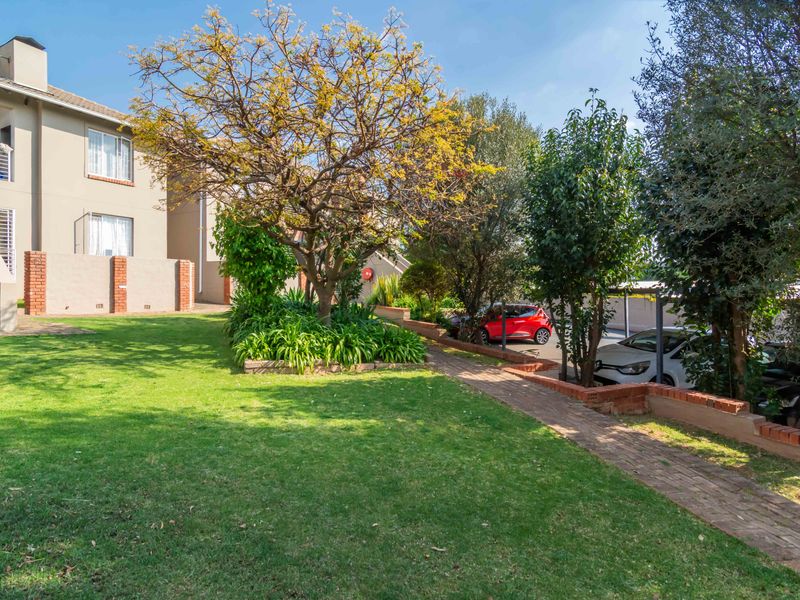 A well-maintained communal garden area featuring a brick pathway, established trees, and a view of the townhouse exterior with balcony access.