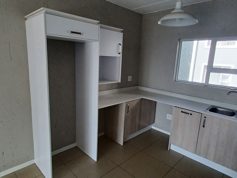 A compact, empty kitchen featuring light wood-finish cabinetry, white countertops, a stainless steel sink, and tiled flooring with an unfinished concrete wall aesthetic.