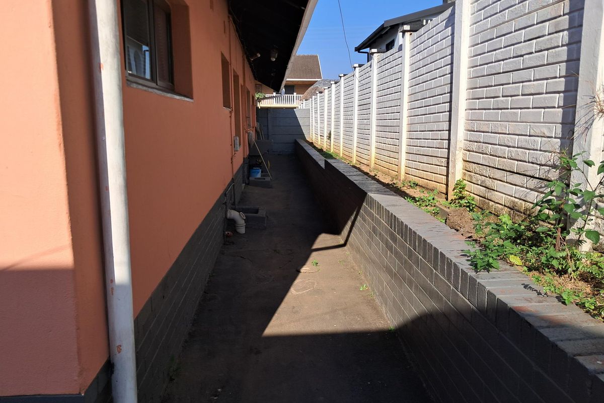 A narrow side passage of the house featuring a concrete boundary wall, a brick retaining wall, and exterior drainage infrastructure.