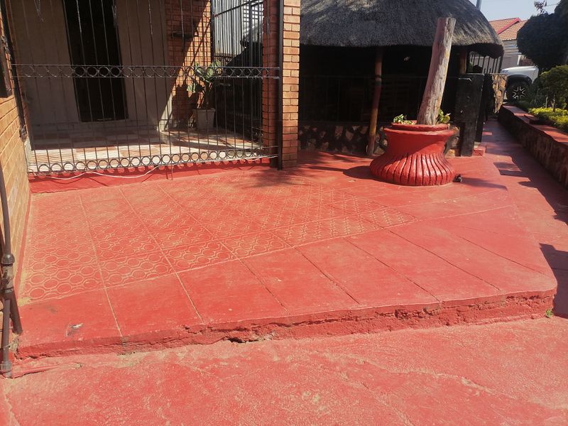 An outdoor patio area featuring painted concrete flooring, brick pillars, and a metal security gate, adjacent to a thatched roof structure.