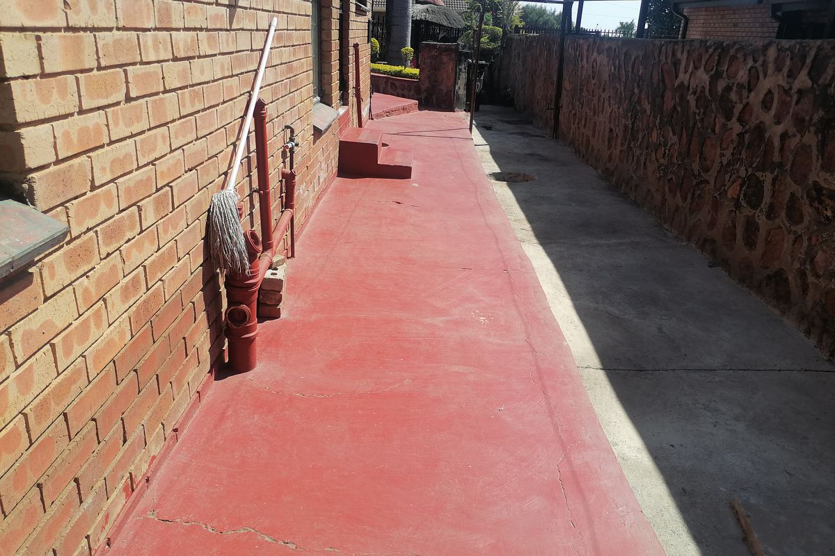 A narrow rear exterior pathway featuring a painted concrete surface, brick walling, and stone-clad boundary walling.