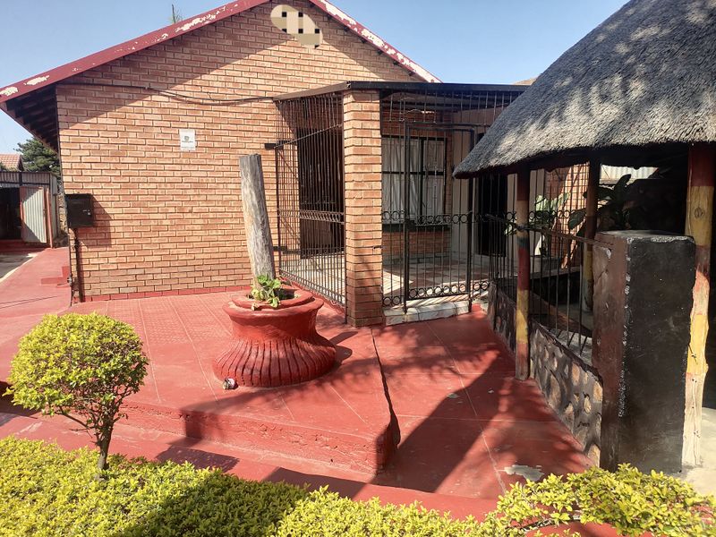 Rear exterior view featuring a brick house facade, a covered patio with security gates, and a traditional thatched lapa structure.