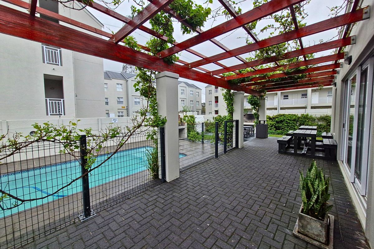 Outdoor pool area featuring a paved patio, timber pergola, and built-in braai, providing a functional communal space for residents.