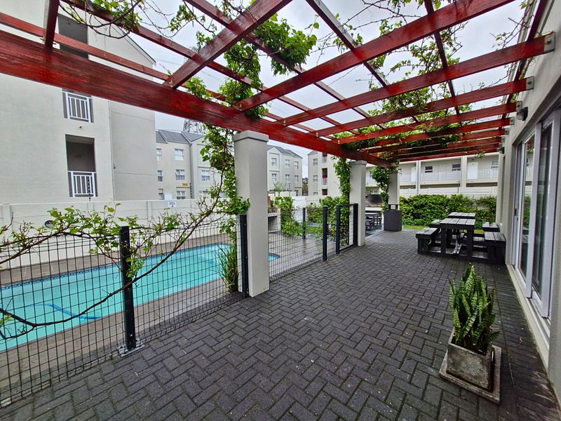 Outdoor pool area featuring a paved patio, timber pergola, and built-in braai, providing a functional communal space for residents.