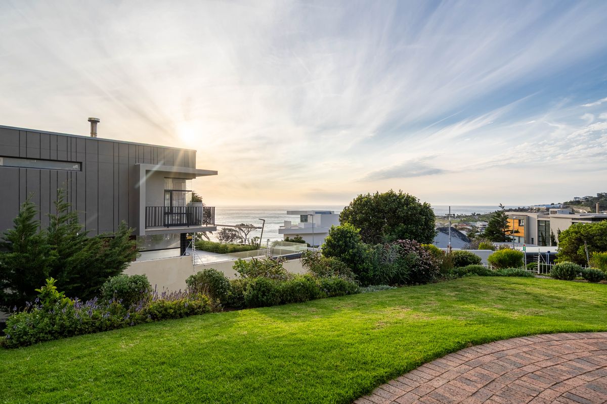 A well-maintained rear garden featuring a brick pathway, lush lawn, and a modern apartment building with a balcony overlooking the ocean.