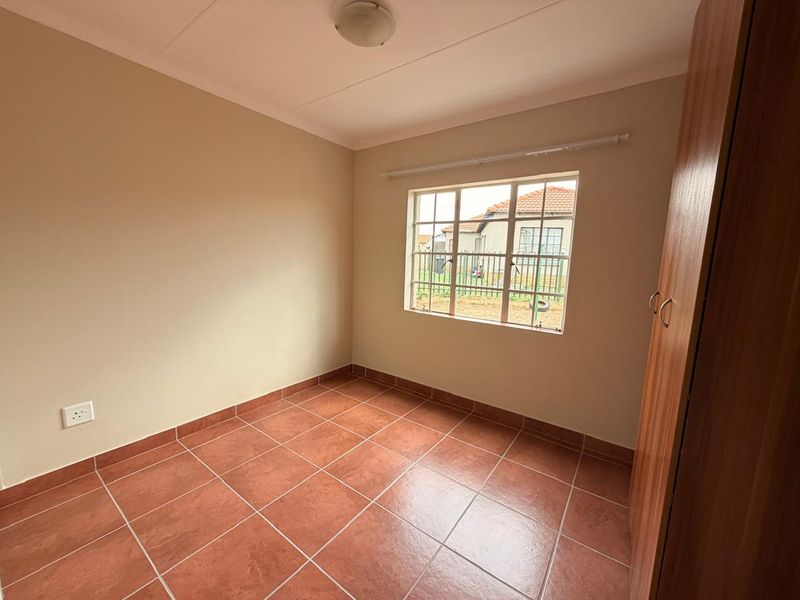 A clean, empty bedroom featuring terracotta-style floor tiles, a built-in cupboard, and a window with security bars.
