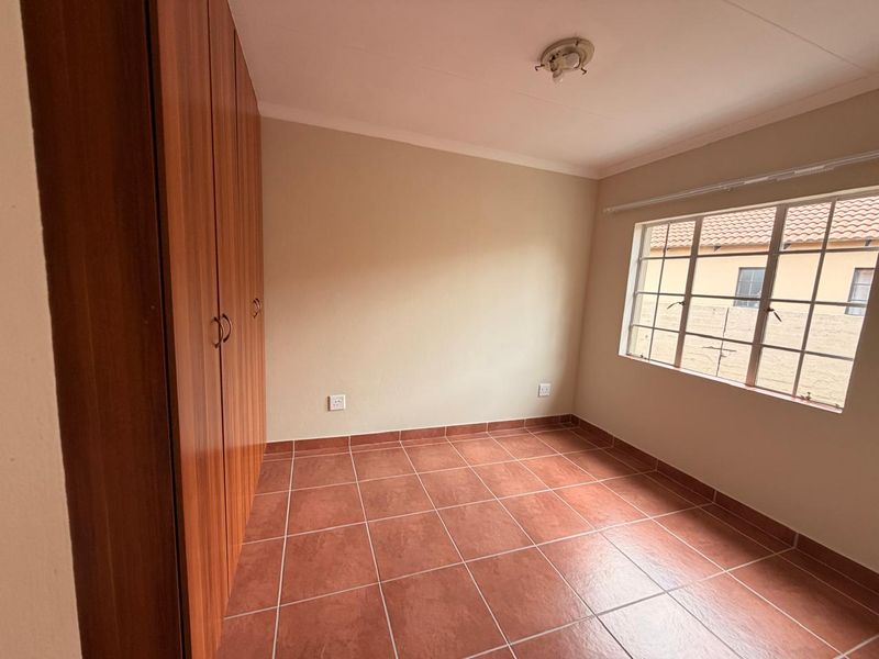 A clean, empty bedroom featuring tiled flooring, built-in wooden cupboards, and a window with security bars.