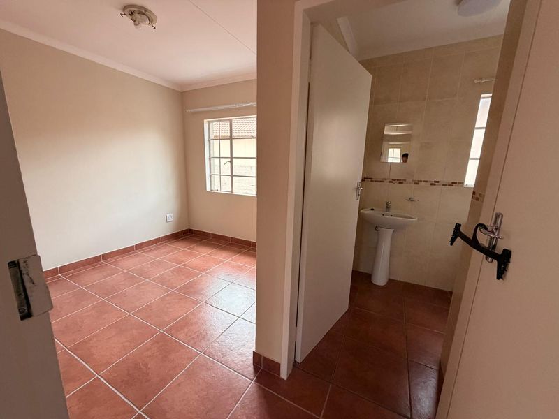 An empty bedroom featuring tiled flooring and an en-suite bathroom area with a pedestal basin and wall-mounted mirror.