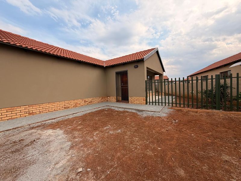 Rear view of the property featuring a terracotta tiled roof, brick plinth detailing, security gate, and a concrete walkway leading to an unlandscaped garden area.