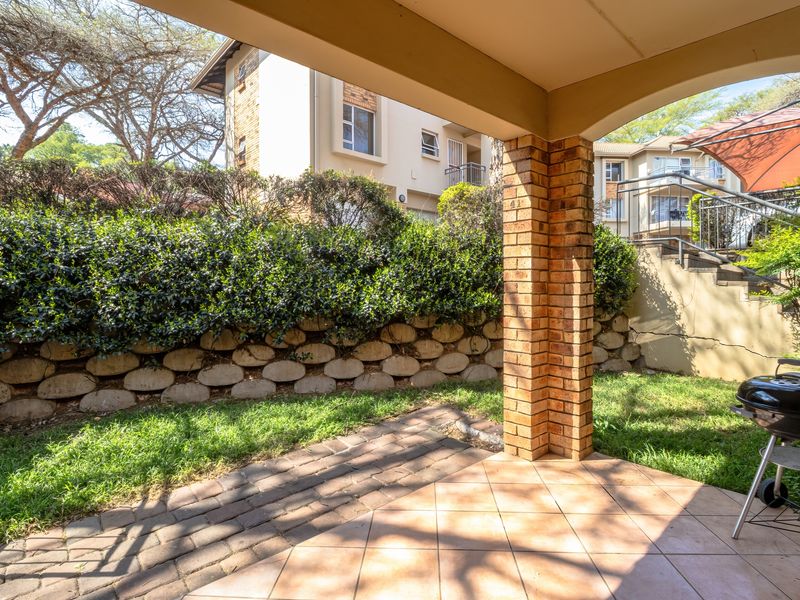 A covered patio area featuring tiled flooring and a brick support pillar, overlooking a small garden with a retaining wall and exterior stairs.