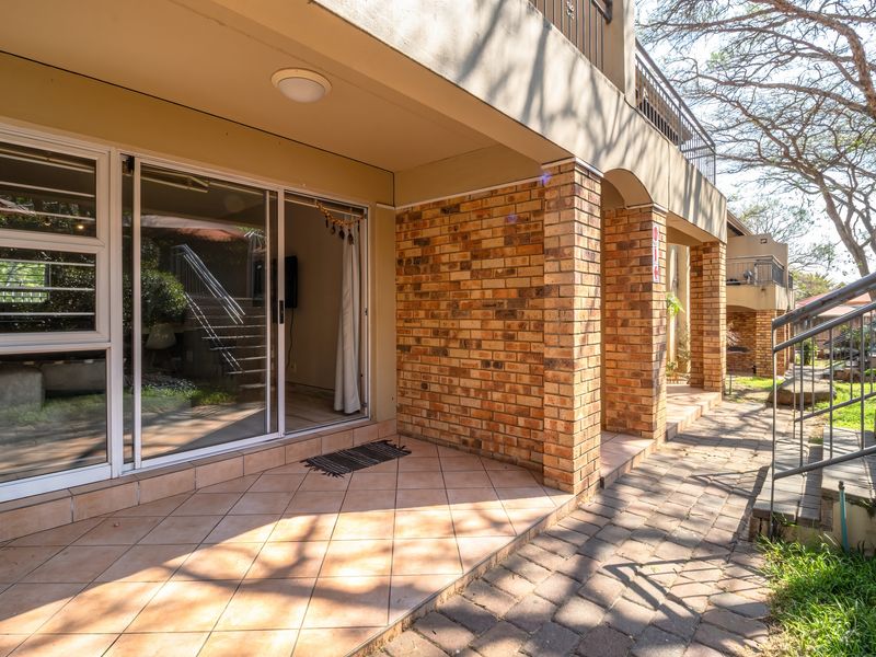 A covered ground-floor patio featuring tiled flooring, brick support pillars, and sliding glass door access to the interior living space.
