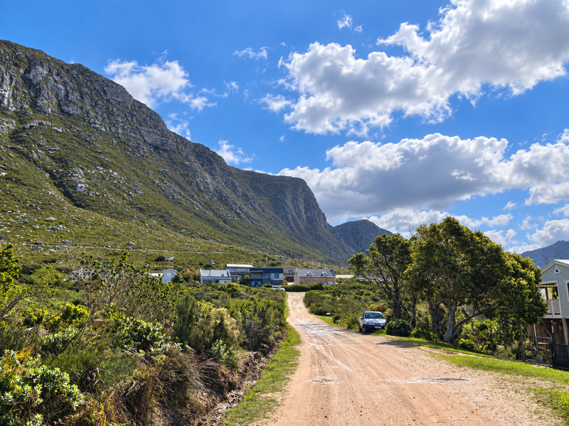 A scenic street view showing an unpaved access road leading towards residential properties nestled against a prominent mountain range.