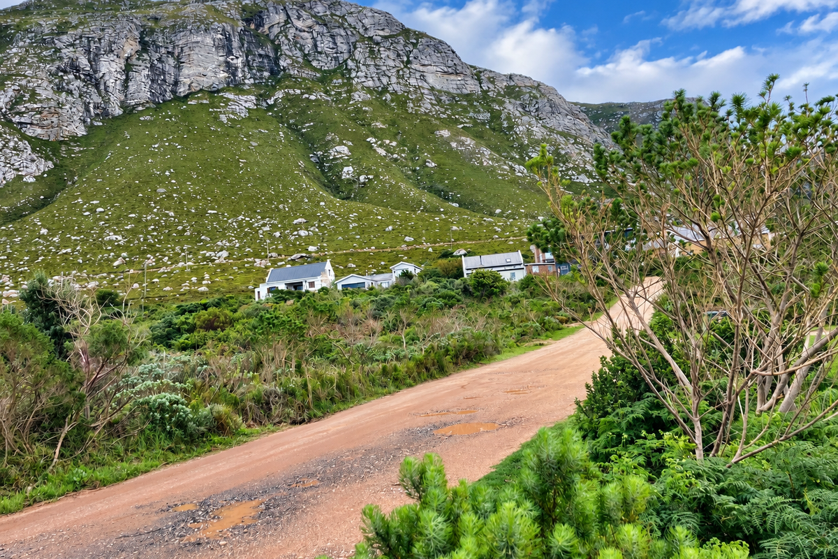 A scenic view of a rural unpaved road leading towards residential properties nestled against a large mountain range.