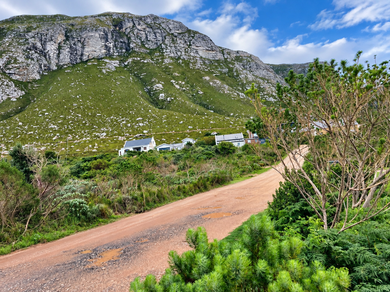 A scenic view of a rural unpaved road leading towards residential properties nestled against a large mountain range.
