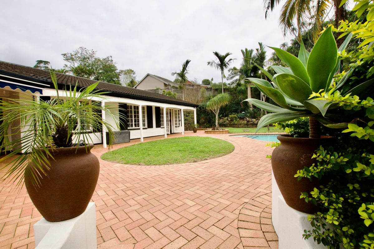 A well-maintained rear exterior featuring a brick-paved patio, covered veranda, swimming pool, and established garden area.