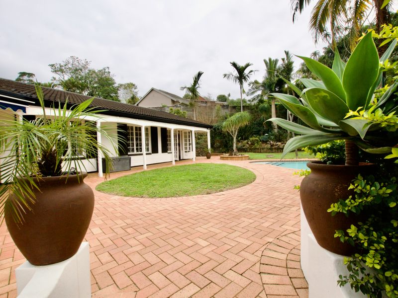 A well-maintained rear exterior featuring a brick-paved patio, covered veranda, swimming pool, and established garden area.