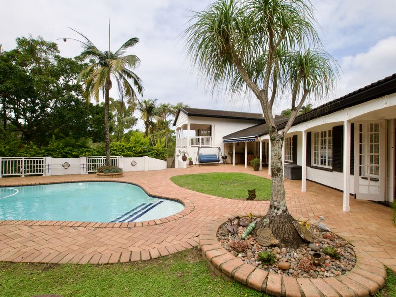 A well-maintained outdoor entertainment area featuring a swimming pool, brick-paved patio, covered veranda, and established garden landscaping.