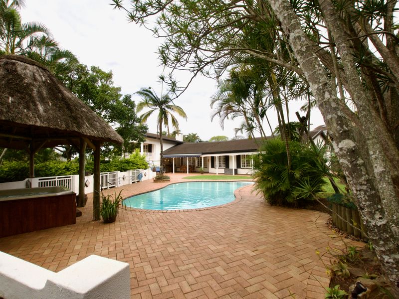 Spacious outdoor pool area featuring a brick-paved patio, a thatched gazebo, and established garden surroundings, ideal for outdoor entertaining.