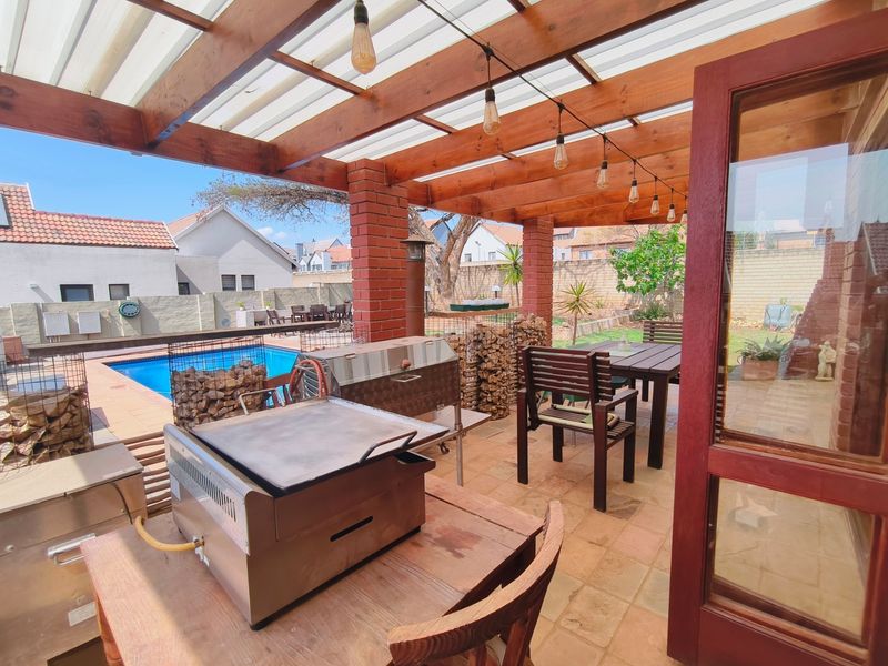 Covered outdoor patio featuring a timber pergola, brick pillars, and built-in braai area, overlooking a private swimming pool and garden.