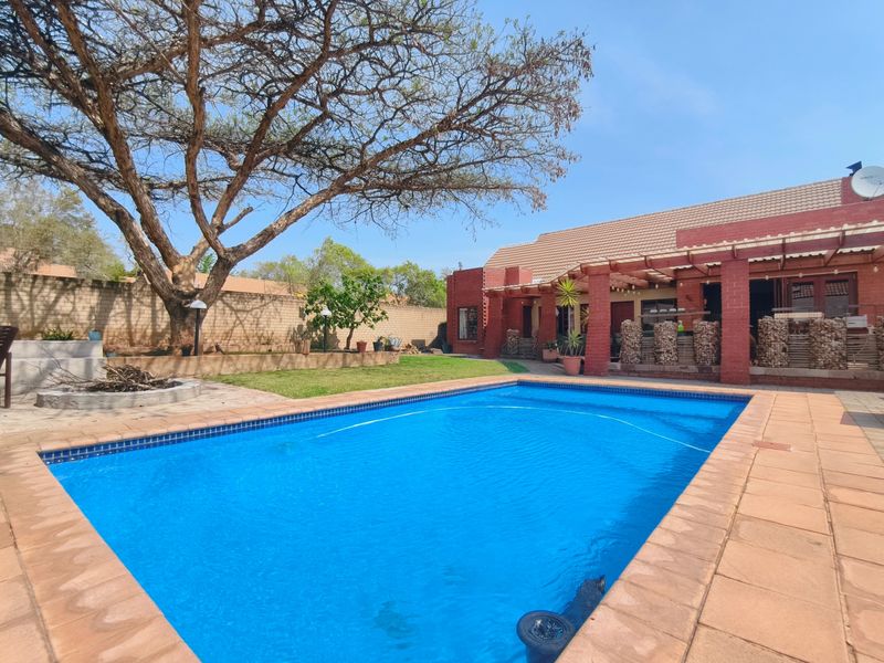 A well-maintained outdoor pool area featuring a large swimming pool, brick-paved patio, and a covered pergola attached to the main house structure.