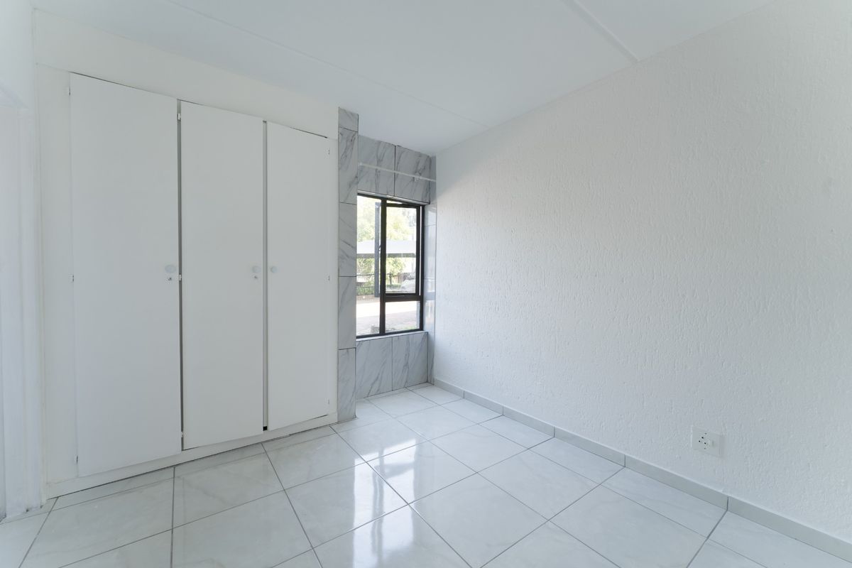 A bright, empty bedroom featuring white built-in cupboards, modern tiled flooring, and a window with marble-look tile detailing.