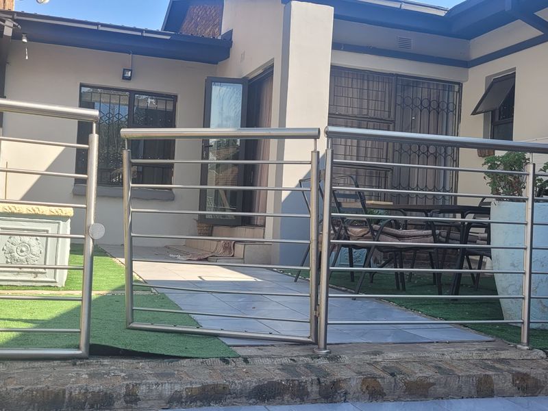 A paved patio area featuring a stainless steel balustrade, artificial grass sections, and direct access to the home through security-fitted doors.