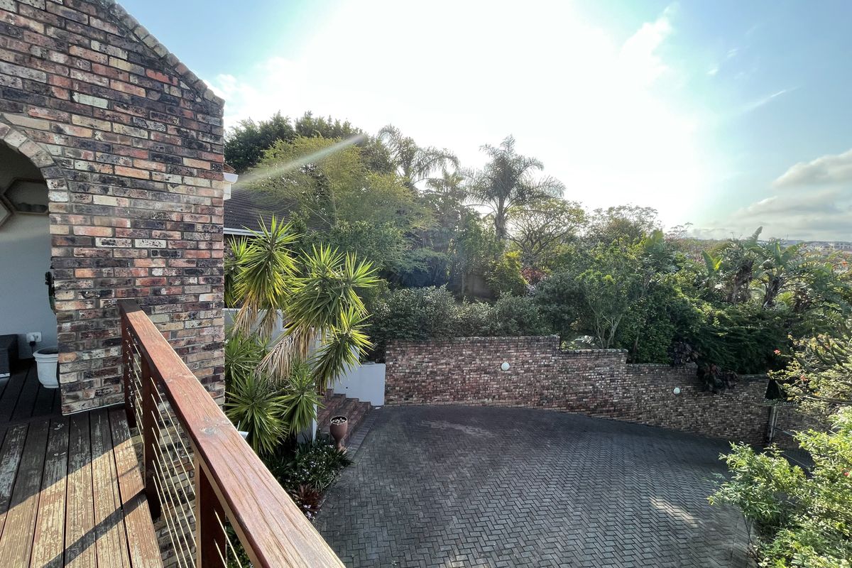An elevated view of a rear outdoor area featuring a timber deck, brick retaining wall, and a paved driveway surrounded by established garden greenery.