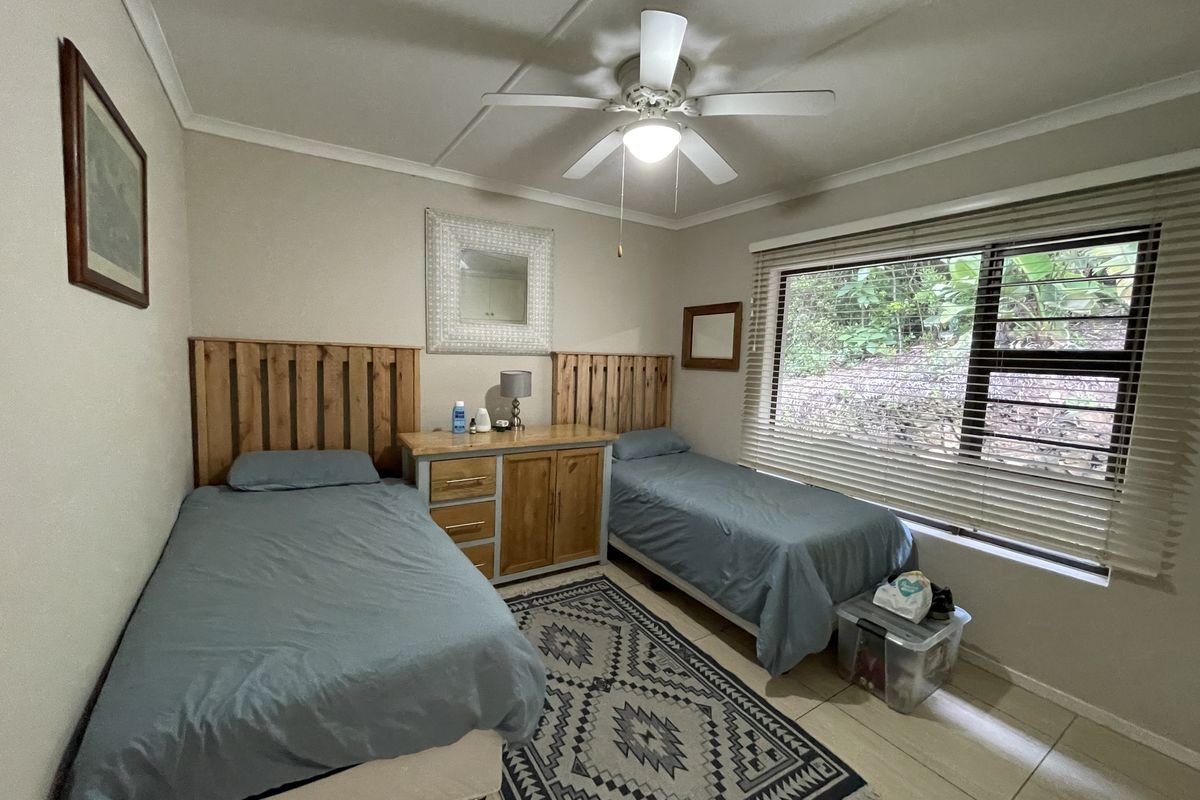 A functional bedroom featuring tiled flooring, a ceiling fan, and large windows with horizontal blinds, providing natural light and a view of the garden.
