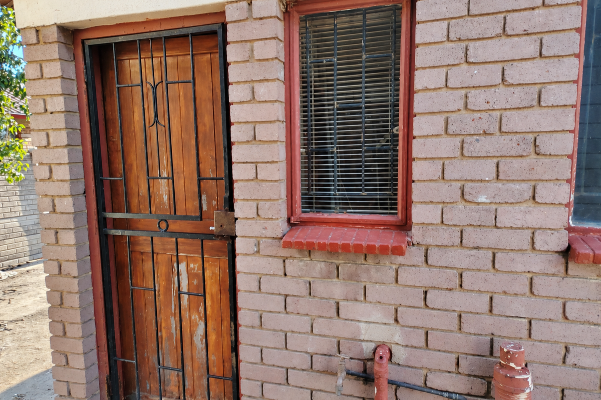 Exterior view of a brick building featuring a wooden door with a security gate, a window with security bars, and external plumbing fixtures.