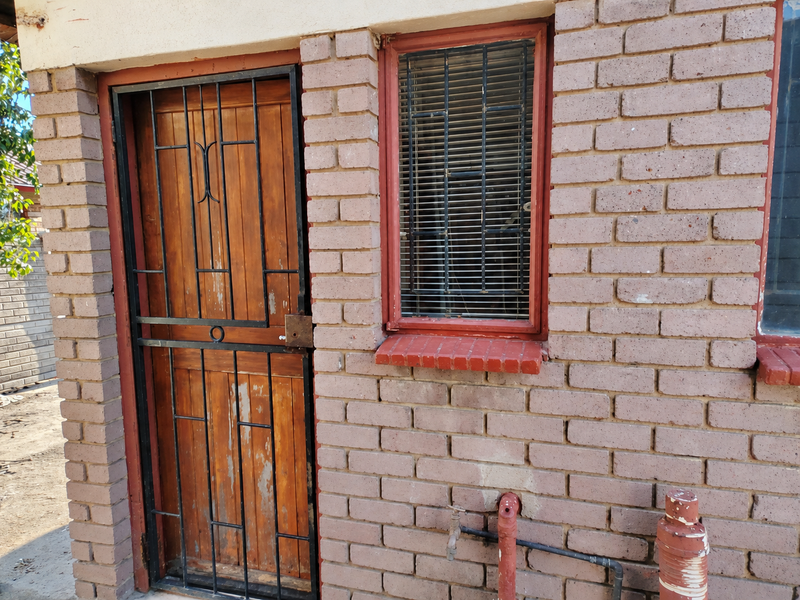 Exterior view of a brick building featuring a wooden door with a security gate, a window with security bars, and external plumbing fixtures.