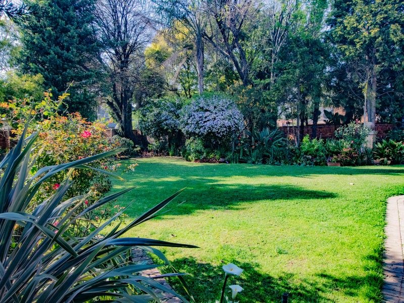 A well-maintained, lush green garden featuring mature trees, established landscaping, and a brick paved pathway leading through the property's rear outdoor space.