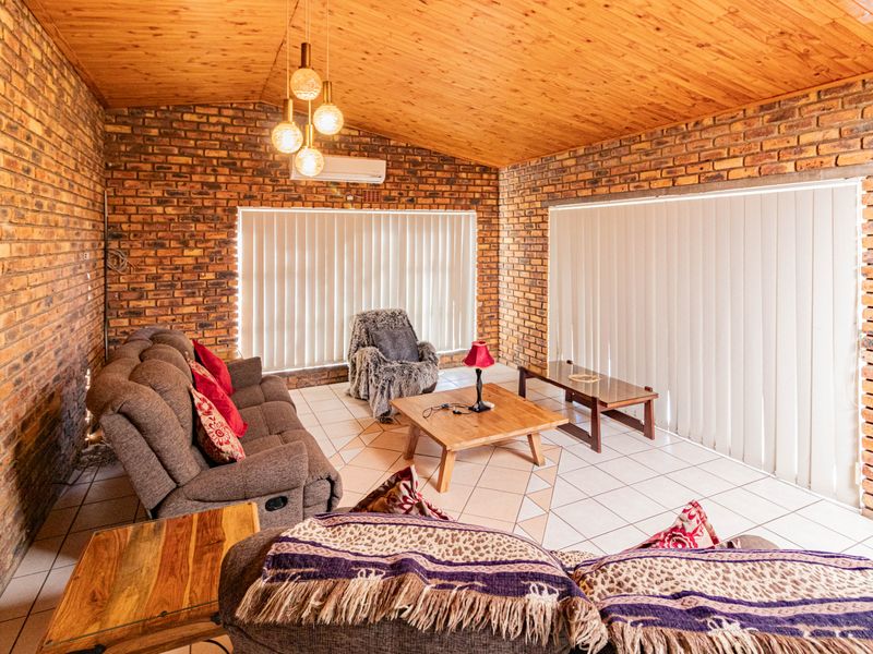 Spacious living room featuring exposed brick walls, a warm timber-paneled ceiling, tiled flooring, and large windows with vertical blinds for natural light.