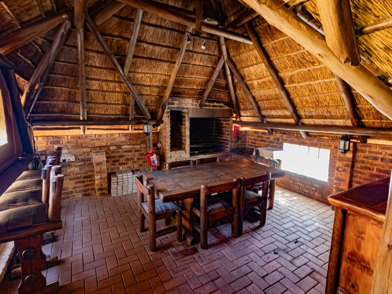 A rustic covered outdoor entertainment area featuring a traditional thatched roof, exposed timber beams, brick walls, and a built-in braai facility.