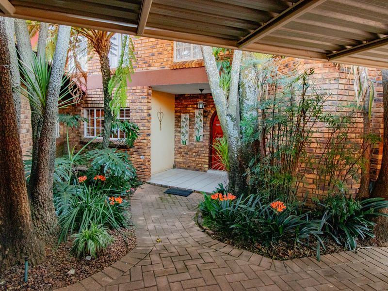 A charming brick-faced front entrance featuring a paved walkway, mature garden landscaping, and a covered carport area.