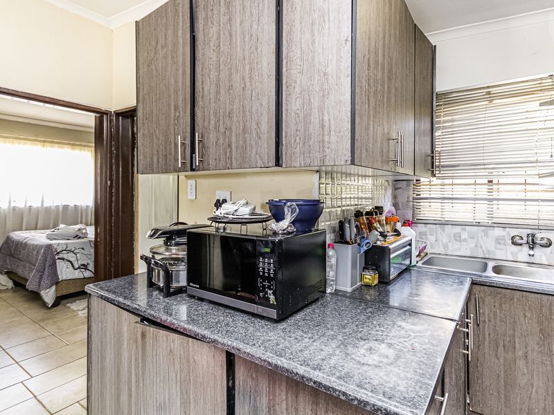 Compact kitchen featuring laminate cabinetry, granite-look countertops, a double bowl stainless steel sink, and tiled backsplash, with a view into the adjacent bedroom.