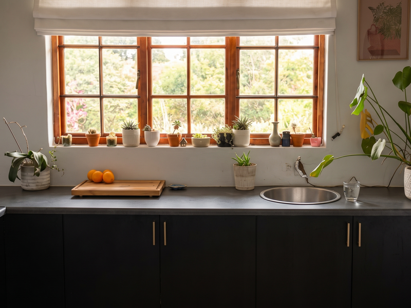 A modern kitchen featuring dark cabinetry, a round stainless steel sink, and a large timber-framed window providing natural light and garden views.