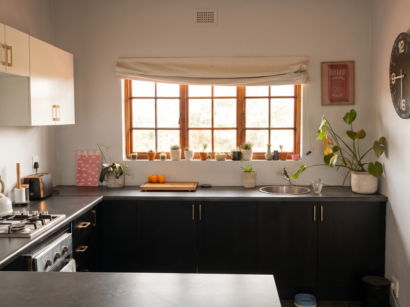 A functional kitchen featuring dark lower cabinetry, white upper cabinets, a gas hob, built-in oven, and a stainless steel sink under a timber-framed window.