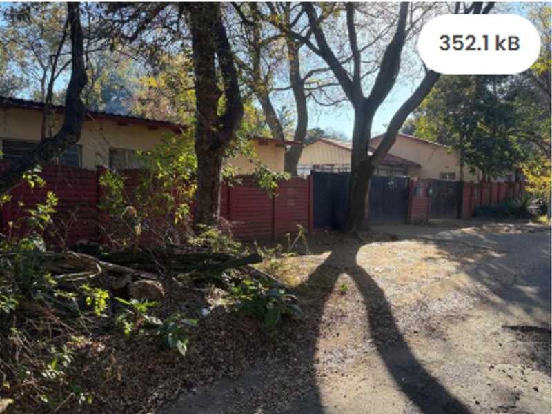 Street view of the property frontage featuring perimeter fencing, a metal gate, and mature trees along the unpaved verge.