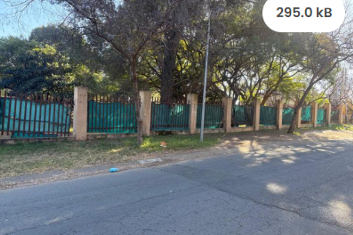 Street view of a vacant residential plot featuring a perimeter wall with metal fencing and mature trees.