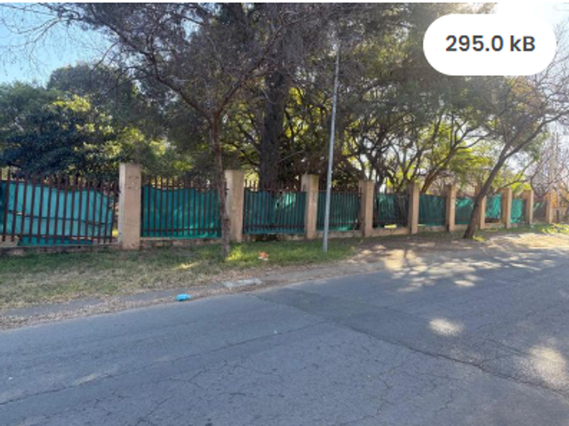 Street view of a vacant residential plot featuring a perimeter wall with metal fencing and mature trees.